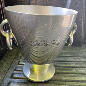 Puede incluir: Una cubeta de champ&aacute;n plateada con forma c&oacute;nica y ancha, con una base circular. Cuenta con dos asas grandes y curvas y las palabras "CHAMPAGNE Nicolas Feuillatte" en escritura elegante. La cubeta tiene un acabado de metal cepillado.