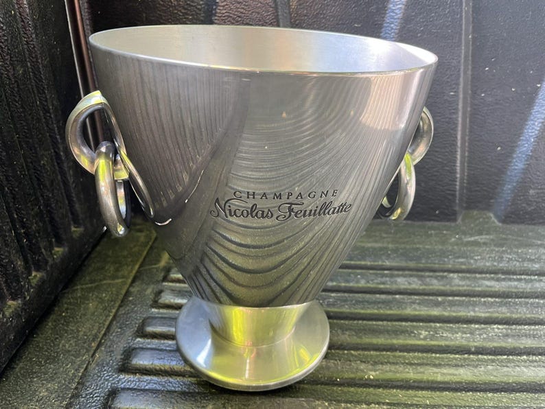 Puede incluir: Una cubeta de champ&aacute;n plateada con forma c&oacute;nica y ancha, con una base circular. Cuenta con dos asas grandes y curvas y las palabras "CHAMPAGNE Nicolas Feuillatte" en escritura elegante. La cubeta tiene un acabado de metal cepillado.