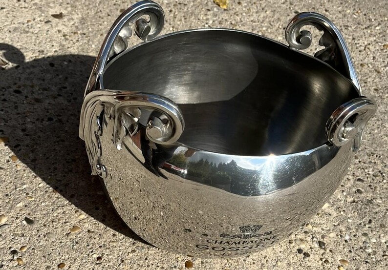 Puede incluir: Una cubitera de champ&aacute;n plateada con asas ornamentadas. La cubitera tiene forma redondeada y superficie reflectante, con las palabras "CHAMPAGNE GOSSET" visibles. Las asas presentan adornos de volutas.
