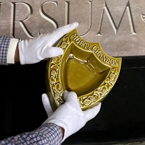 K&ouml;nnte beinhalten: Eine schildf&ouml;rmige, olivgr&uuml;ne Keramikschale mit dem goldenen Schriftzug "MO&Euml;T & CHANDON". Die Schale wird von behandschuhten H&auml;nden gehalten. Der Rand ist mit einem Weinrebenmuster verziert.
