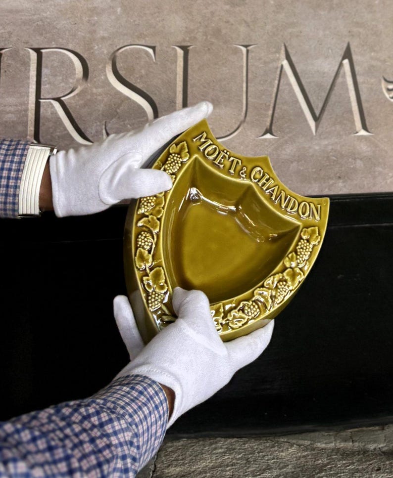 K&ouml;nnte beinhalten: Eine schildf&ouml;rmige, olivgr&uuml;ne Keramikschale mit dem goldenen Schriftzug "MO&Euml;T & CHANDON". Die Schale wird von behandschuhten H&auml;nden gehalten. Der Rand ist mit einem Weinrebenmuster verziert.