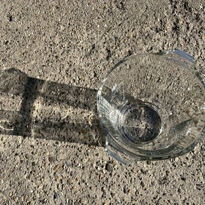 May include: A clear glass bowl with a round base and two handles. The bowl is on a rough, grey surface, and its shadow is cast to the left. The bowl appears to be empty.