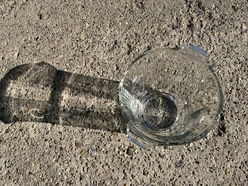 May include: A clear glass bowl with a round base and two handles. The bowl is on a rough, grey surface, and its shadow is cast to the left. The bowl appears to be empty.