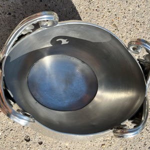 Puede incluir: Un cuenco de metal plateado con asas decorativas. El cuenco tiene forma redondeada con una amplia abertura y un interior liso y reflectante. Las asas son curvas y presentan detalles en forma de hojas. El cuenco es probablemente para fines decorativos o de servicio.