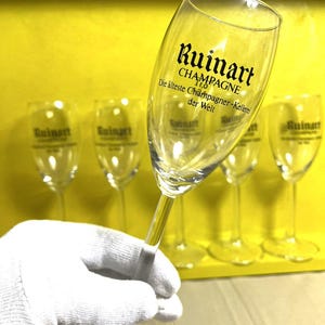 May include: A clear champagne flute with the "Ruinart" logo and text. The text reads "CHAMPAGNE" and "Die &auml;lteste Champagner-Kellerei der Welt". The flute is held by a gloved hand, with other flutes in the background.
