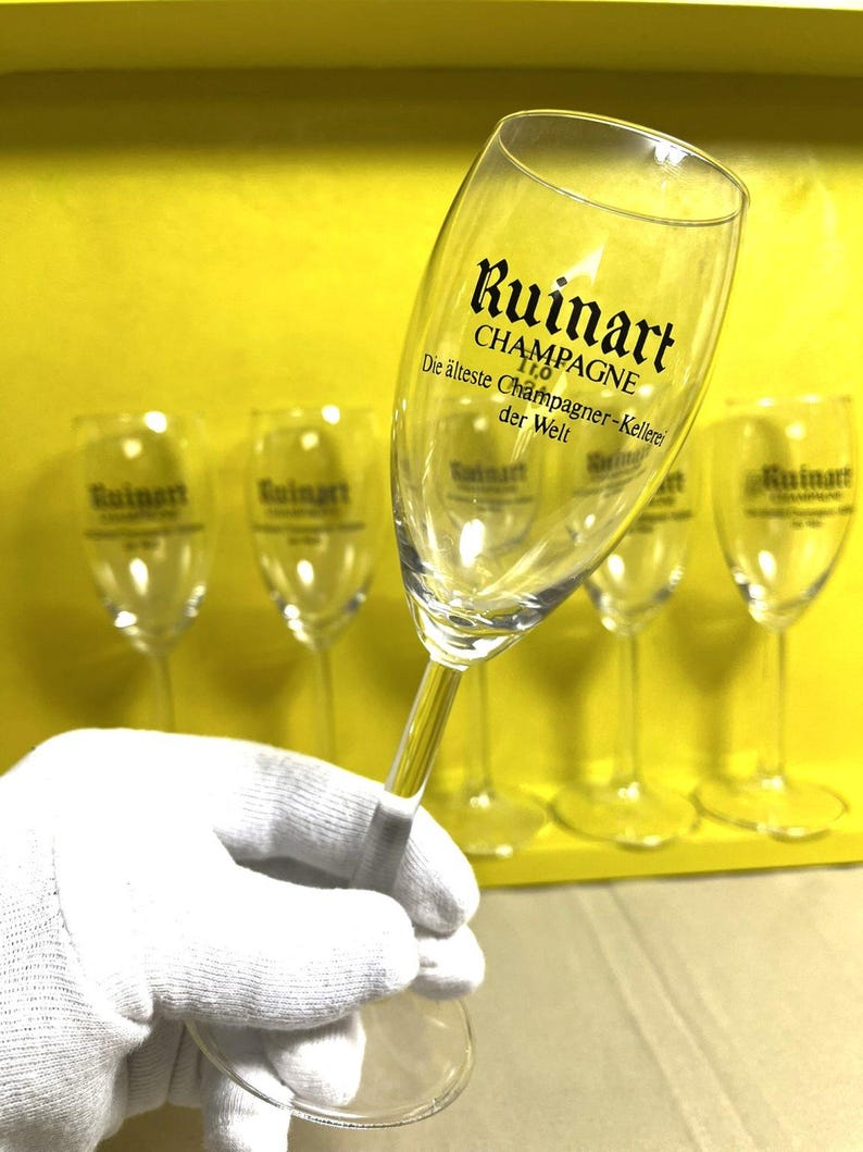 May include: A clear champagne flute with the "Ruinart" logo and text. The text reads "CHAMPAGNE" and "Die &auml;lteste Champagner-Kellerei der Welt". The flute is held by a gloved hand, with other flutes in the background.