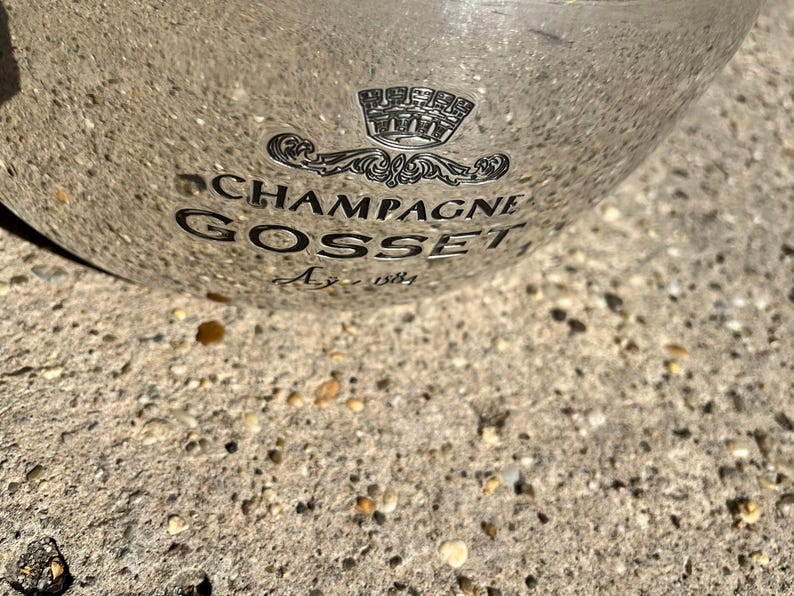 Puede incluir: Una cubeta de champ&aacute;n plateada con las palabras "CHAMPAGNE GOSSET Ay - 1584" impresas en negro. La cubeta tiene un emblema decorativo encima del texto. La cubeta est&aacute; sobre una superficie rugosa y texturizada.