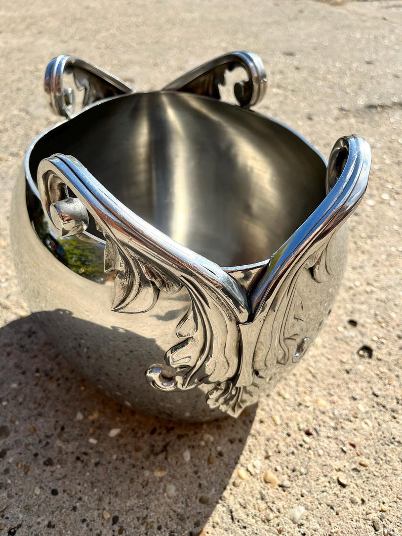 Puede incluir: Un cubo de hielo de metal plateado pulido con asas ornamentadas y detalles decorativos de hojas. El cubo tiene un cuerpo redondeado y una amplia abertura. Las asas se curvan hacia arriba y hacia afuera, lo que se suma al dise&ntilde;o elegante. Para enfriar bebidas.