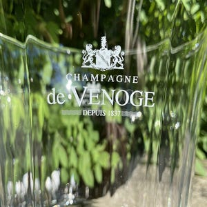 May include: A clear glass champagne bucket with a fluted design. The bucket features the text "CHAMPAGNE de VENOGE DEPUIS 1837" in white lettering, along with a crest. The background is blurred green foliage.
