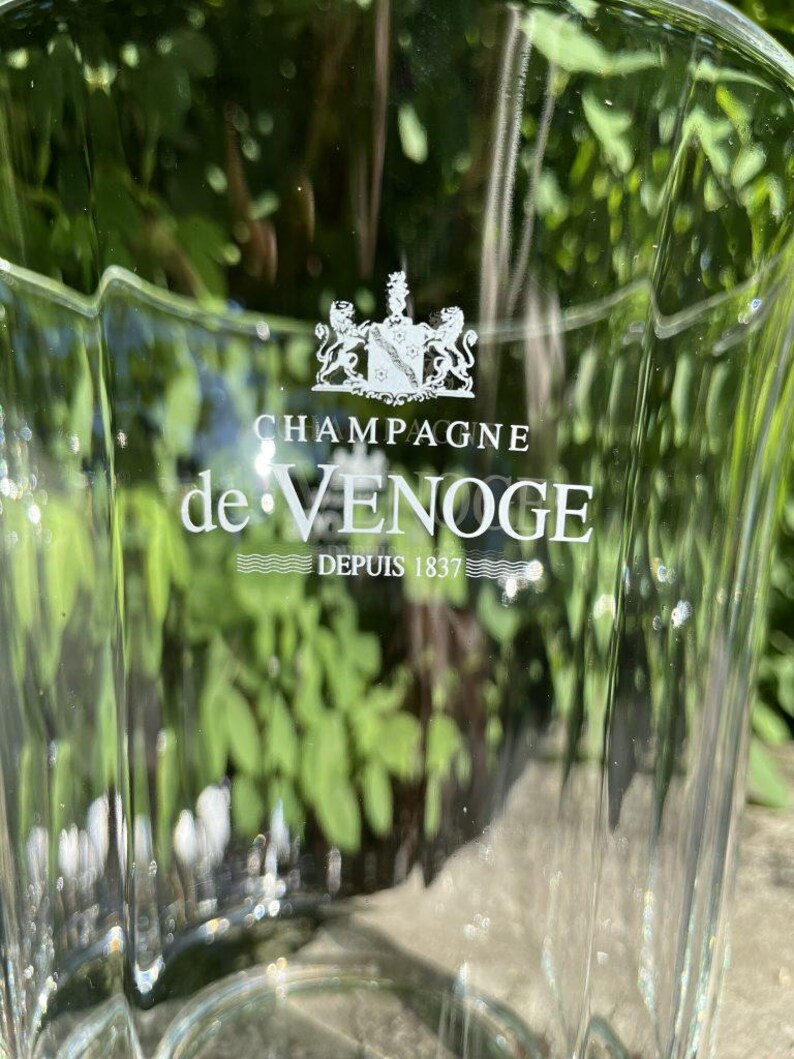 May include: A clear glass champagne bucket with a fluted design. The bucket features the text "CHAMPAGNE de VENOGE DEPUIS 1837" in white lettering, along with a crest. The background is blurred green foliage.