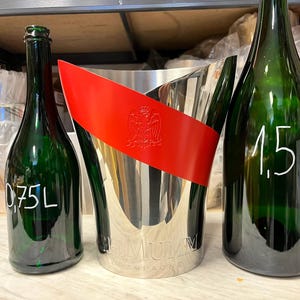 May include: Three green champagne bottles, one labeled 0.75L and another 1.5L, flank a silver champagne bucket with a red band. The bucket has the word "G.H. MUMM" on it.