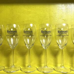 May include: Six clear champagne flutes with the "Ruinart Champagne" logo printed on the front. The glasses are arranged in a row against a yellow background. The text "Champagne" is visible on the glasses.