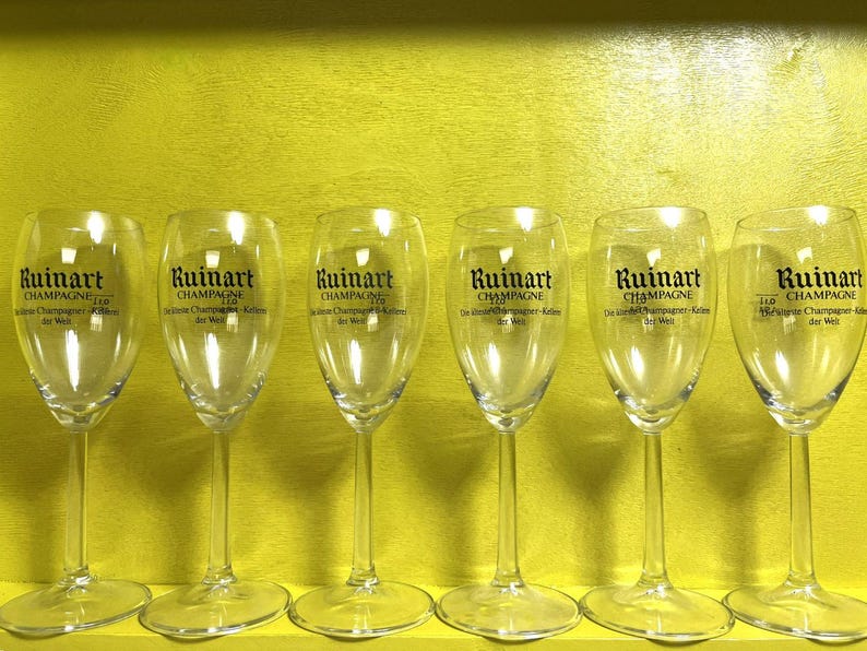 May include: Six clear champagne flutes with the "Ruinart Champagne" logo printed on the front. The glasses are arranged in a row against a yellow background. The text "Champagne" is visible on the glasses.