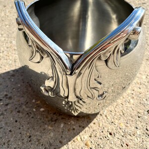 Puede incluir: Cubeta de hielo de metal plateado con detalles ornamentales de hojas y volutas. La cubeta tiene un cuerpo redondeado y asas decorativas. El dise&ntilde;o presenta un patr&oacute;n sim&eacute;trico de hojas y vides.