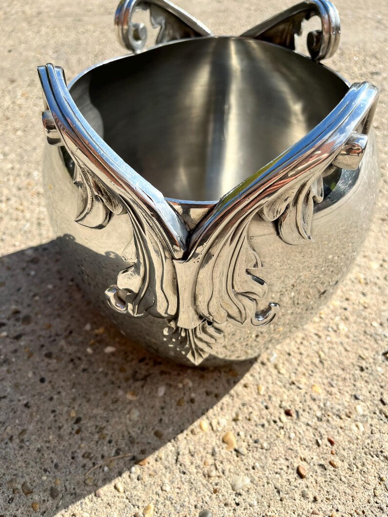 Puede incluir: Cubeta de hielo de metal plateado con detalles ornamentales de hojas y volutas. La cubeta tiene un cuerpo redondeado y asas decorativas. El dise&ntilde;o presenta un patr&oacute;n sim&eacute;trico de hojas y vides.