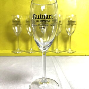 May include: Clear champagne flute with a long stem and a round base. The glass features the "Ruinart CHAMPAGNE" logo in black lettering. Several similar flutes are visible in the background, set against a yellow backdrop.