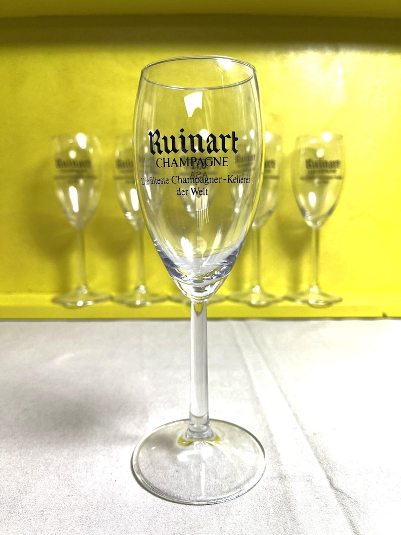 May include: Clear champagne flute with a long stem and a round base. The glass features the "Ruinart CHAMPAGNE" logo in black lettering. Several similar flutes are visible in the background, set against a yellow backdrop.