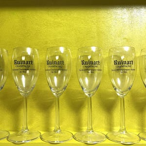 May include: Six clear champagne flutes with the "Ruinart Champagne" logo printed in black. The glasses are arranged in a row against a yellow background. The text "Champagne" is visible on each glass.