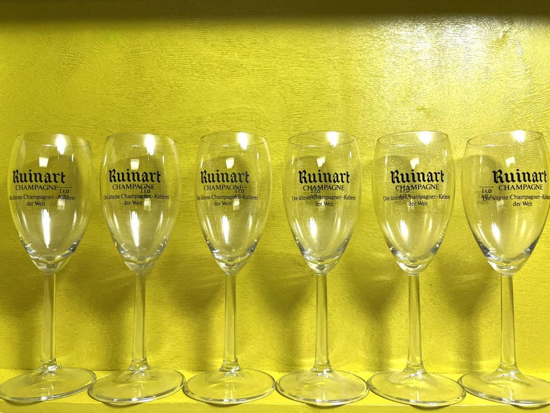 May include: Six clear champagne flutes with the "Ruinart Champagne" logo printed in black. The glasses are arranged in a row against a yellow background. The text "Champagne" is visible on each glass.