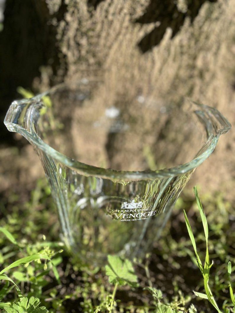 May include: A clear glass champagne bucket with a flared shape and two handles. The bucket has the words "Champagne VENOGE" printed on the front. The bucket is sitting on a bed of green grass.