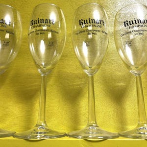May include: Clear champagne flutes with long stems are arranged in a row. The flutes have a rounded bowl shape and the text "Ruinart Champagne" is printed on the glass. The flutes are on a yellow surface.