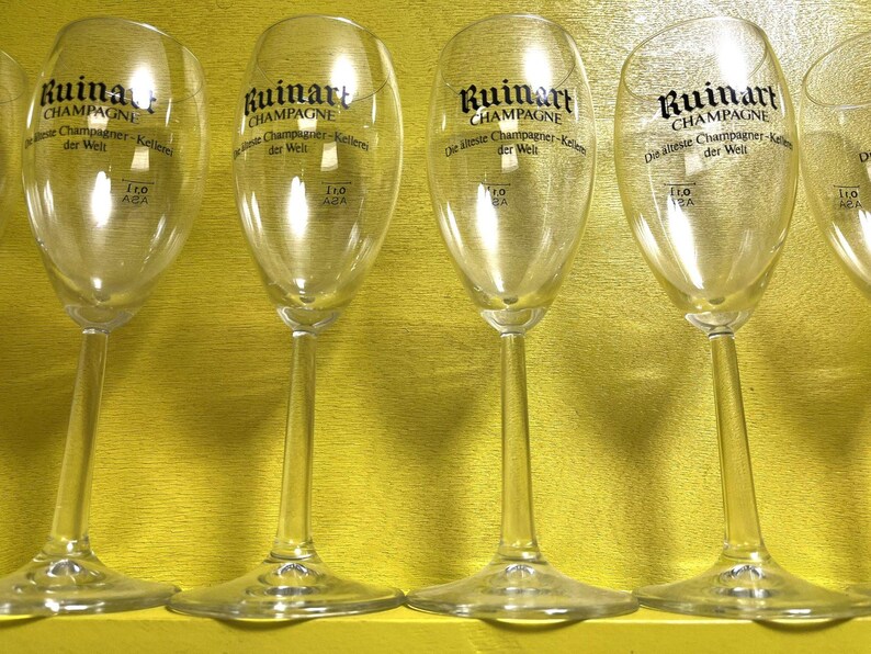 May include: Clear champagne flutes with long stems are arranged in a row. The flutes have a rounded bowl shape and the text "Ruinart Champagne" is printed on the glass. The flutes are on a yellow surface.