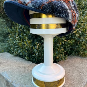 May include: A blue and brown tweed newsboy cap with a black brim, displayed on a white stand with a gold band.