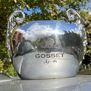 Puede incluir: Cubitera de champ&aacute;n plateada con asas ornamentadas. La cubitera est&aacute; grabada con las palabras "CHAMPAGNE GOSSET Ay, 1584" y un emblema de corona. La superficie brillante refleja el entorno.