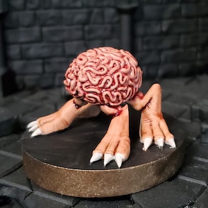 Op de afbeelding: Een miniatuurfiguur van een brein met benen en klauwen, geschilderd in tinten roze en rood, staand op een zwarte en gouden basis.