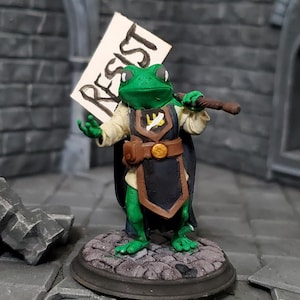 May include: A green frog figurine holding a sign that reads "RESIST". The frog is wearing a black cape, a brown belt, and a cream-colored shirt. The figurine is standing on a stone base.