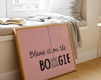 May include: A square framed wall art piece with a light pink background. The text reads "Blame it on the BOOGIE" in black lettering. A black line hangs from the letter O in the word on, ending in a circular disco ball graphic with star accents.