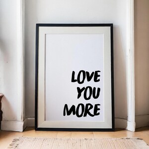 May include: A rectangular wall art print in a black frame with a white mat. The print features the black text LOVE YOU MORE in a bold, hand-lettered style font centered on a plain white background. The frame has a clean, simple design.