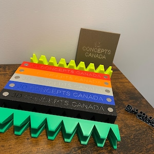 Puede incluir: Un conjunto de seis bloques impresos en 3D de colores vivos con el texto "3D CONCEPTS CANADA" en cada bloque. Los bloques están apilados uno encima del otro y dispuestos en forma escalonada. Los bloques están hechos de un plástico liso y brillante. Los bloques son de color amarillo brillante, naranja, rojo, gris claro, azul y negro.