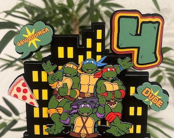 May include: A Teenage Mutant Ninja Turtles themed cake topper with four turtles, a pizza slice, a city skyline, and the words "Cowabunga", "Dude", and "Hayes".