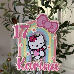 May include: A colorful birthday cake topper featuring Hello Kitty, a rainbow, and the number 17. The name "Karina" is displayed in pink with gold glitter accents. Small white daisies are scattered around the rainbow.