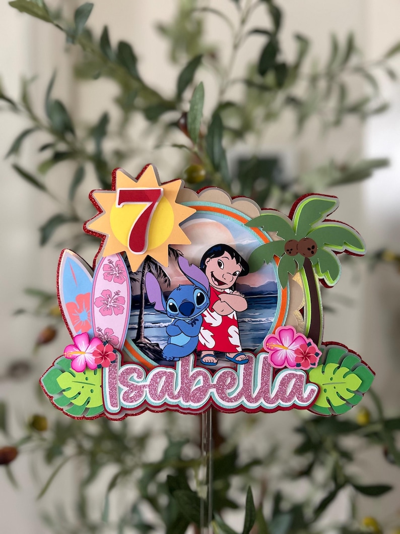 Może przedstawiać: Kolorowa dekoracja na tort z postaciami Lilo i Stitch, deską surfingową i palmą. Numer 7 jest wyświetlany w słońcu, a imię "Isabella" jest napisane r&oacute;żowym brokatem.