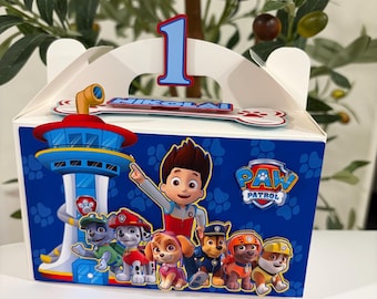 May include: A white party favor box with a blue Paw Patrol design. The box features the Paw Patrol characters, a tower, and the number 1. The name "NIKOLAI" is printed on the box.