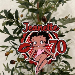 May include: A red and white cake topper with a cartoon image of Betty Boop. The topper says "Jeanette 70" in red glitter letters.