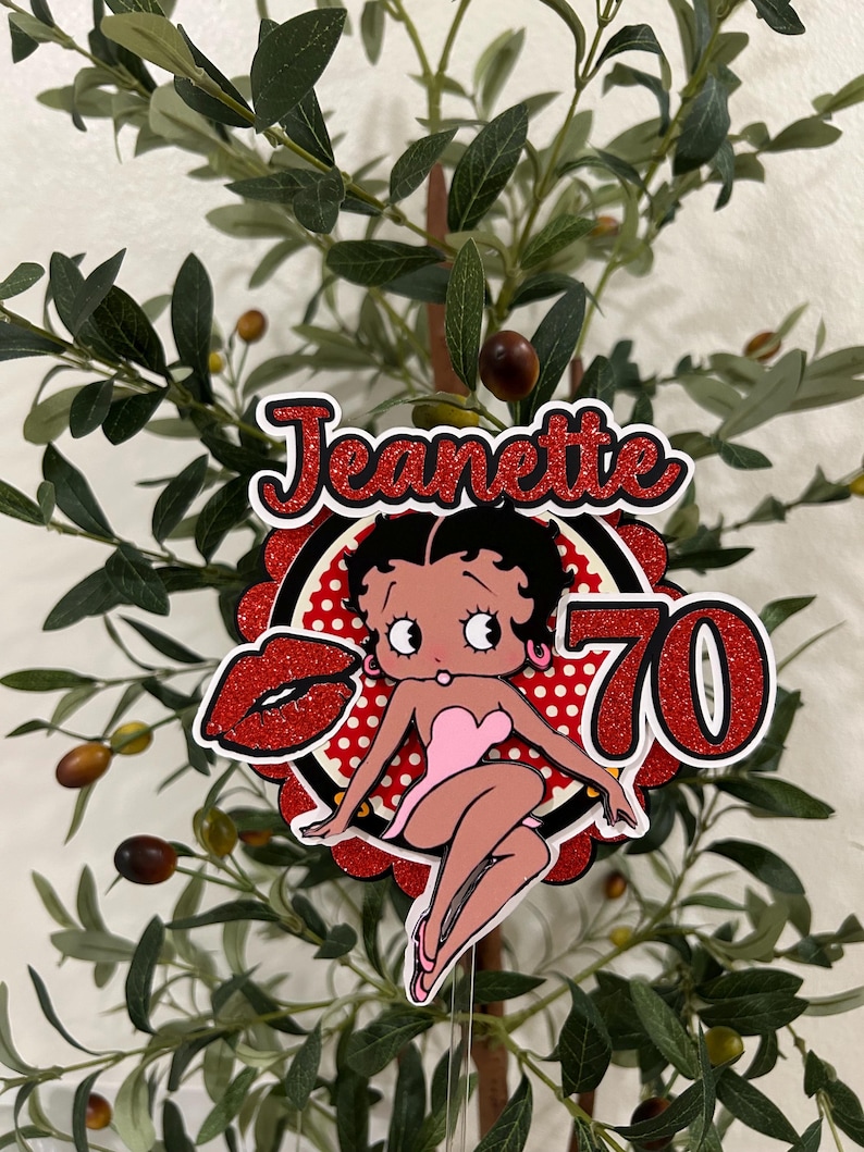 May include: A red and white cake topper with a cartoon image of Betty Boop. The topper says "Jeanette 70" in red glitter letters.