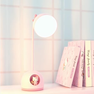 May include: A pink desk lamp with a round, illuminated light. The base features a Hello Kitty figure in a circular window. Pink books with Hello Kitty designs are stacked nearby, creating a cute and functional desk accessory.