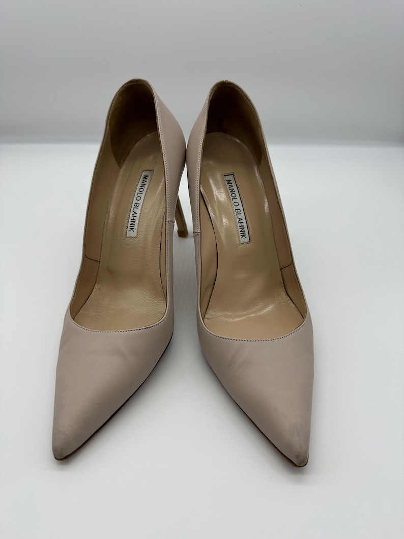 May include: A pair of light beige Manolo Blahnik high heels. The shoes have pointed toes and stiletto heels. The brand name is printed on the insole of each shoe. The heels are approximately 4 inches tall.
