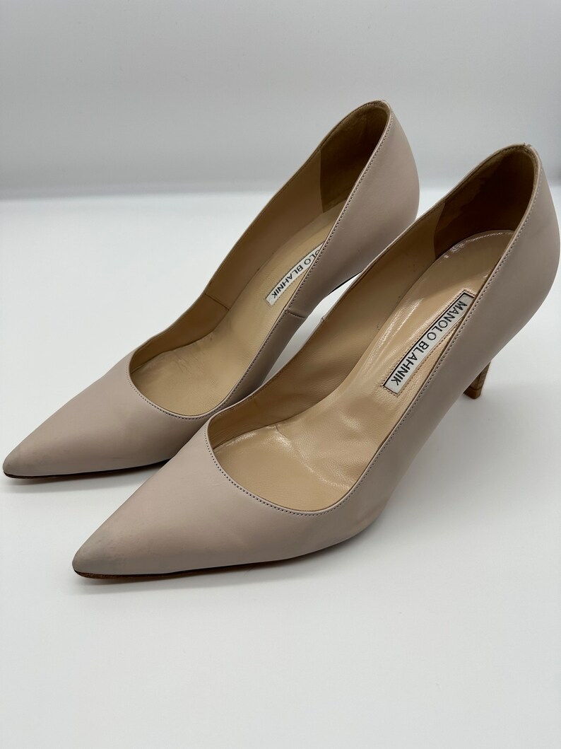 May include: A pair of light beige Manolo Blahnik pumps. The shoes have pointed toes and stiletto heels. The brand name is printed on the insole. The pumps are made of a smooth leather-like material.
