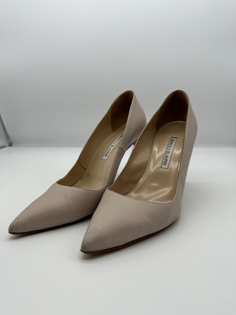 May include: A pair of light beige high-heeled pumps with pointed toes. The shoes have a stiletto heel and a smooth leather finish. The brand name "MANOLO BLAHNIK" is visible on the insole.