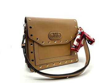 May include: A brown leather shoulder bag with gold studs and a small pocket on the front. The bag has a black strap and a red and white patterned scarf attached to it. The bag has the logo "LOVE MOSCHINO" on the front.