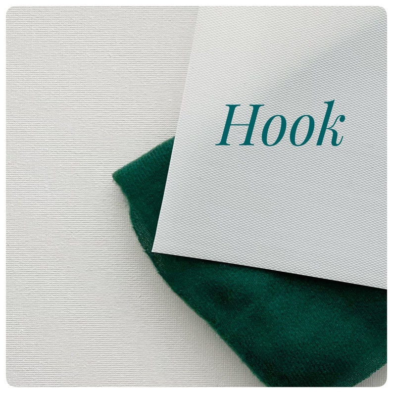May include: A white card with the word "Hook" written in teal blue on a white background. A piece of dark green fabric is partially covering the card.