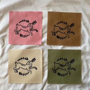 May include: Four square fabric patches in pink, brown, beige, and green. Each patch features a black illustration of a running hare encircled by a leafy wreath. The patches are arranged on a white fabric surface.