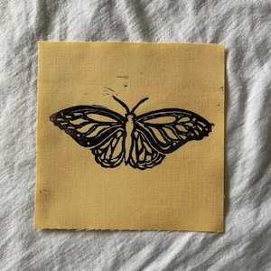 May include: A square, yellow fabric patch featuring a black butterfly print. The butterfly design is detailed with visible wing patterns. The fabric has a slightly textured appearance, and the edges are unfinished.