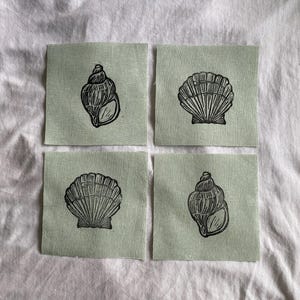 May include: Four square fabric patches in a light sage green color, each featuring a black ink block print of a seashell. Two patches display a spiral-shaped shell, while the other two patches display a fan-shaped scallop shell design.