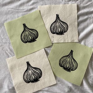 May include: Four fabric squares with a black garlic bulb print. Two squares are a light green color, and two are off-white. The garlic bulb design is a detailed linocut style, with visible lines and textures. The squares are arranged on a white, textured surface.
