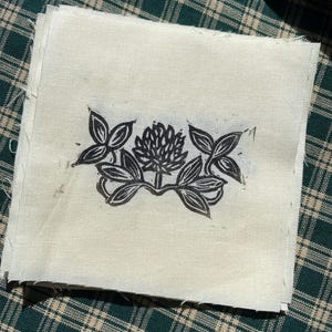 May include: A black and white linocut print of a clover flower on a white fabric background.